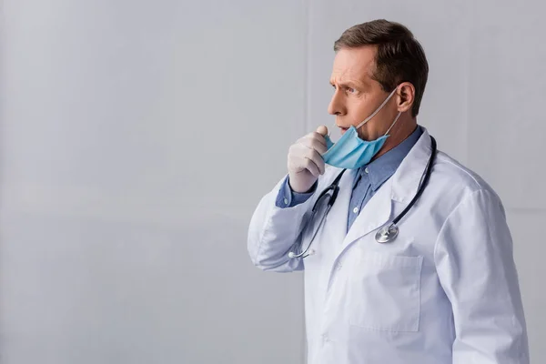 Зрілий лікар в медичній масці кашляє на сірому — стокове фото