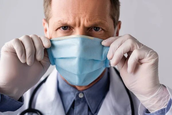 Mature doctor touching blue medical mask isolated on grey — Stock Photo