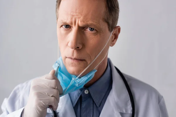 Doctor maduro en guante de látex tocando máscara médica azul sobre gris - foto de stock