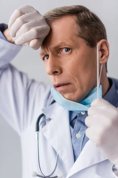 Tired mature doctor in latex gloves touching blue medical mask isolated on grey — Stock Photo