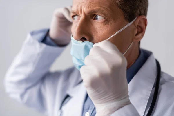 Селективное внимание врача средних лет в латексных перчатках, касающихся синей медицинской маски, изолированной на сером — стоковое фото