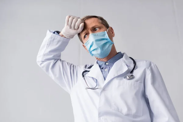 Tired and mature doctor in medical mask and latex gloves touching forehead on grey — Stock Photo