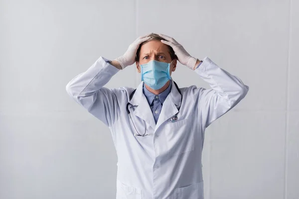 Stressato medico di mezza età in maschera medica blu toccare la testa sul grigio — Foto stock