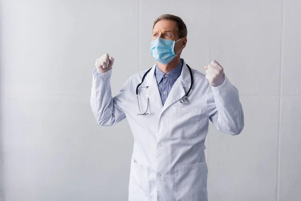 Напружений лікар середнього віку в блакитній медичній масці, що стоїть з зіпсованими кулаками на сірому — стокове фото