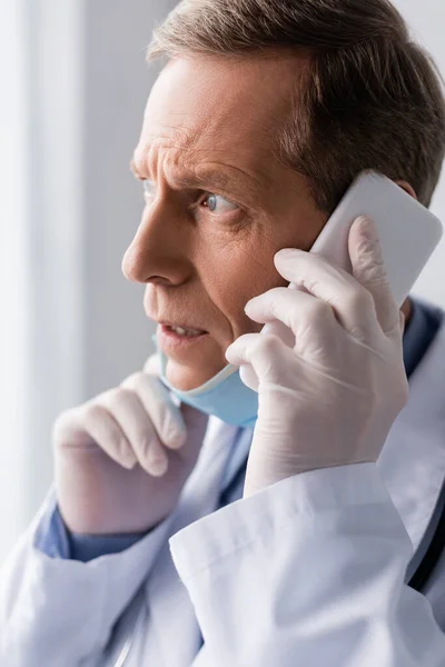 Stressed and mature doctor in white coat talking on smartphone — Stock Photo