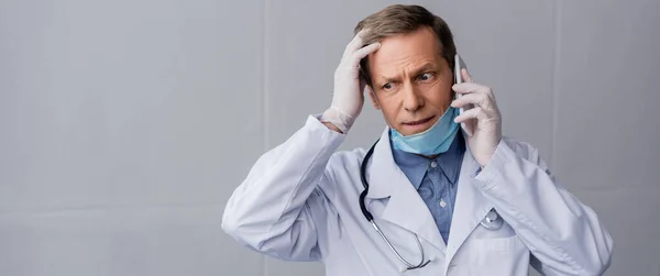 Tiro panorâmico de médico de meia-idade estressado falando no smartphone em cinza — Fotografia de Stock