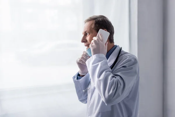 Glücklicher und reifer Arzt im weißen Kittel, der am Fenster mit dem Smartphone spricht — Stockfoto