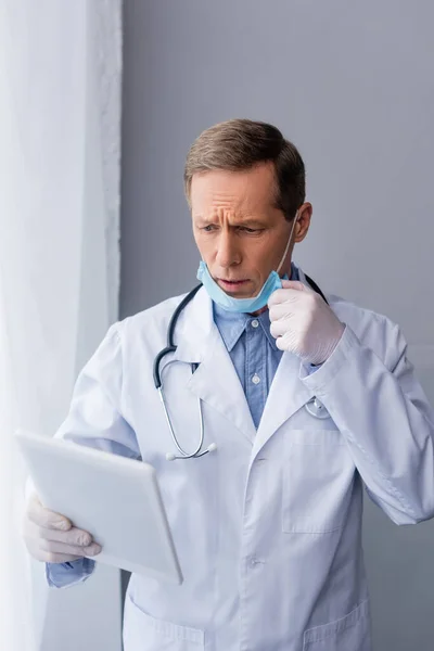Besorgter reifer Arzt in medizinischer Maske blickt auf digitales Tablet auf grau — Stockfoto