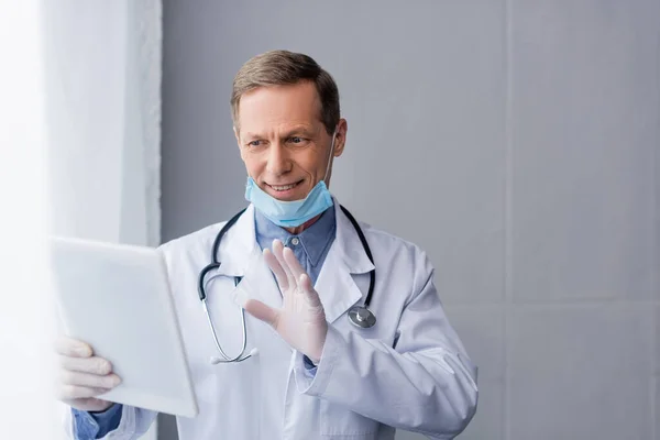 Heureux mature médecin dans masque médical ayant appel vidéo sur gris — Photo de stock