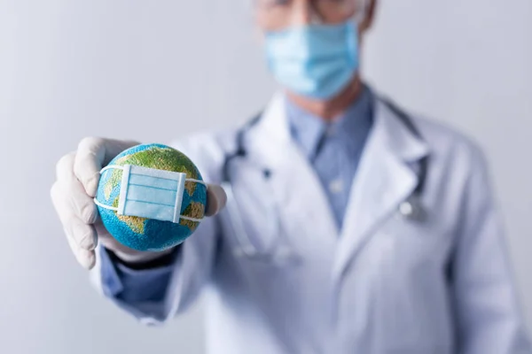 Selective focus of mature doctor in latex glove holding small globe in medical mask on grey — Stock Photo