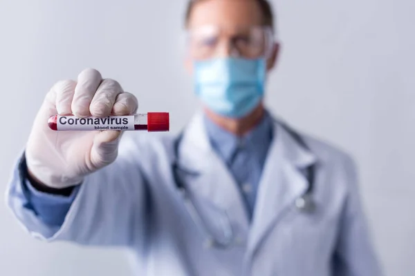 Selektiver Fokus des reifen Arztes in medizinischer Maske im Reagenzglas mit Schriftzug der Coronavirus-Blutprobe isoliert auf grau — Stockfoto