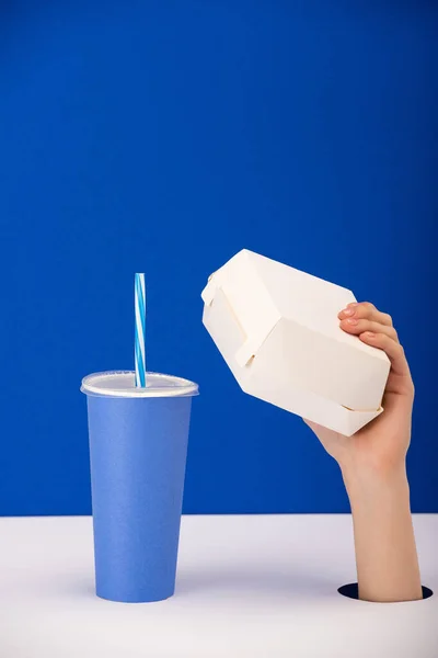 Vista ritagliata della donna che tiene scatola di cartone vicino alla tazza di carta con bibita isolata su blu — Foto stock