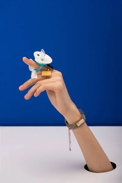 Cropped view of woman with bracelet and rings holding silicone pacifier isolated on blue — Stock Photo