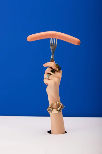 Vue recadrée de la femme avec bracelet et anneaux tenant fourchette avec saucisse isolée sur bleu — Photo de stock