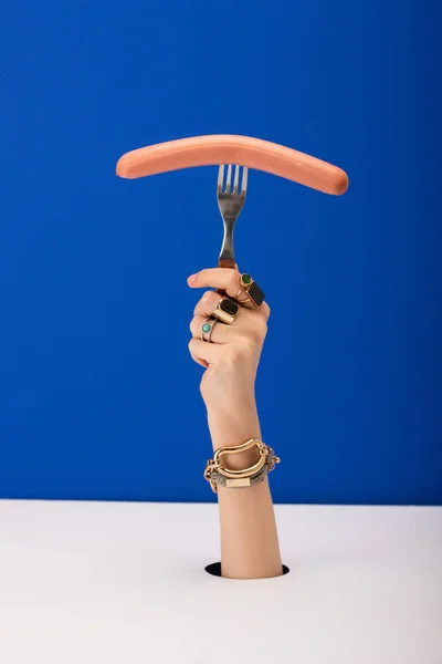 Vista recortada de mujer con brazalete y anillos sosteniendo tenedor con salchicha aislada en azul - foto de stock