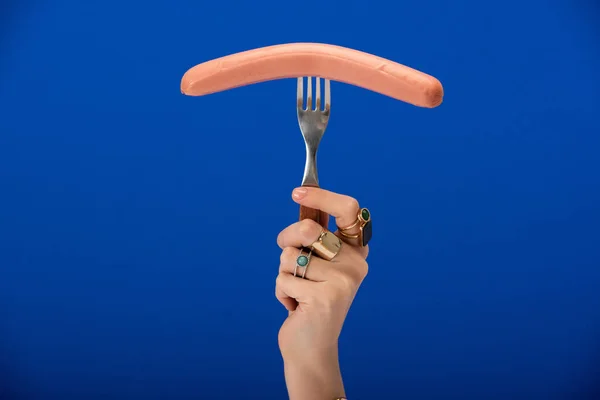 Cropped view of woman with rings on fingers holding fork with sausage on blue — Stock Photo