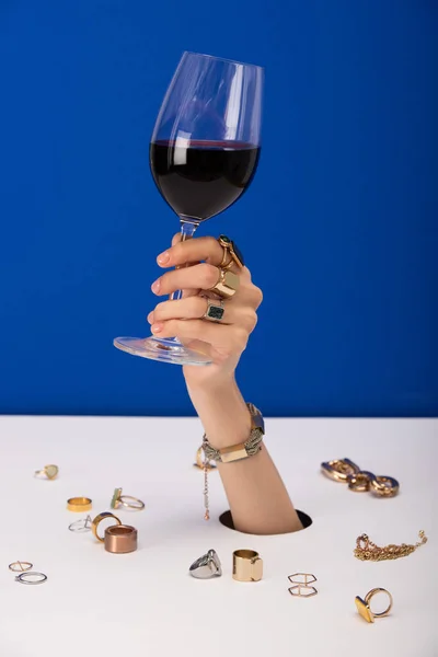 Vista recortada de la mujer con brazalete en la mano celebración de vidrio con vino tinto cerca de anillos aislados en azul - foto de stock