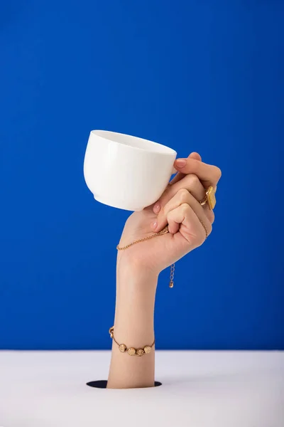 Cropped view of woman with bracelet on hand holding cup of coffee isolated on blue — Stock Photo