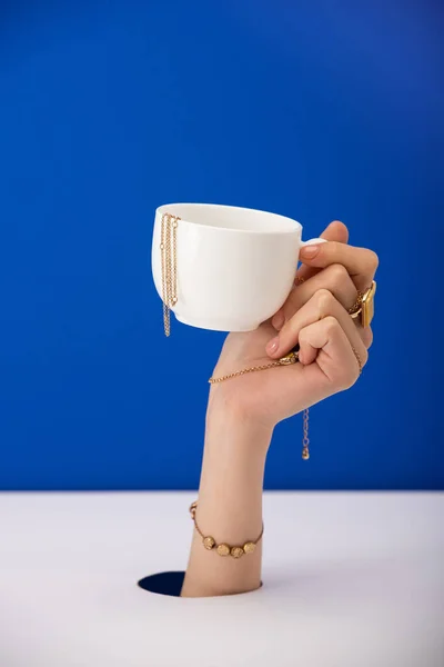 Vista ritagliata della donna con bracciale sulla tazza mano con catena d'oro isolato su blu — Foto stock