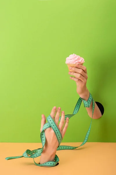 Cropped view of woman with measuring tape on hands holding cupcake on green and orange — Stock Photo