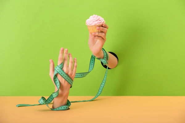 Vue recadrée de la femme avec ruban à mesurer sur les mains tenant délicieux cupcake sur vert et orange — Photo de stock