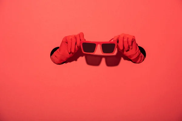 Обрезанный вид женщины в перчатках, держащей стильные солнечные очки на красном — стоковое фото
