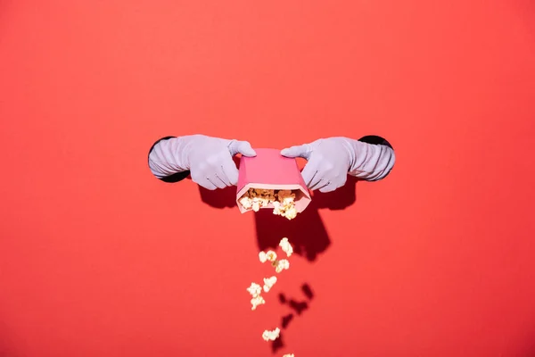 Ausgeschnittene Ansicht einer Frau in Handschuhen, die Popcorn aus einem Eimer auf Rot gießt — Stockfoto