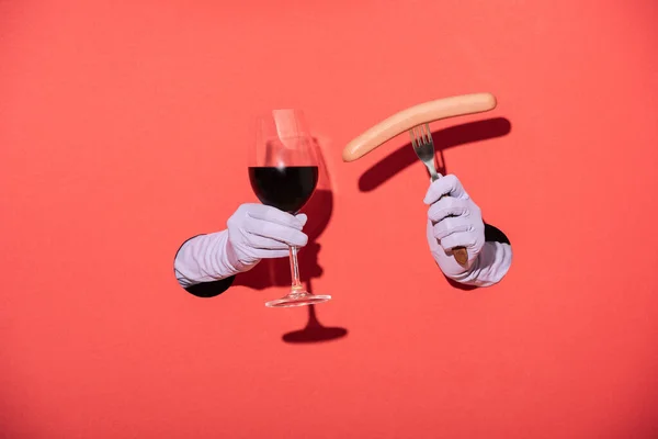 Vue recadrée de la femme dans des gants tenant verre avec vin rouge et fourchette avec saucisse sur rouge — Photo de stock