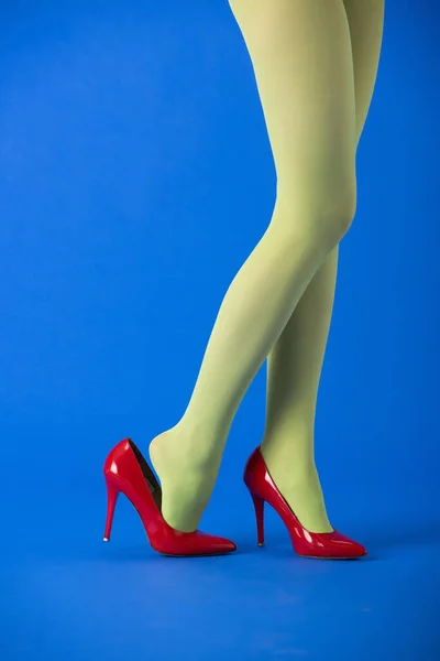 Vista cortada de modelo elegante em meias verdes e saltos vermelhos em pé no azul — Fotografia de Stock