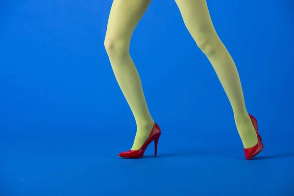 Vue recadrée du modèle en collants lumineux et talons rouges posant sur bleu — Photo de stock