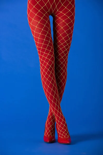 Cropped view of model in fishnet tights and red heels standing with crossed legs on blue — Stock Photo