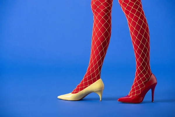 Vista recortada de la mujer en medias de rejilla, tacones amarillos y rojos de pie sobre azul - foto de stock