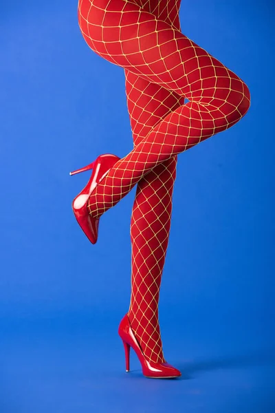 Cropped view of stylish woman in fishnet tights and red heels posing on blue — Stock Photo