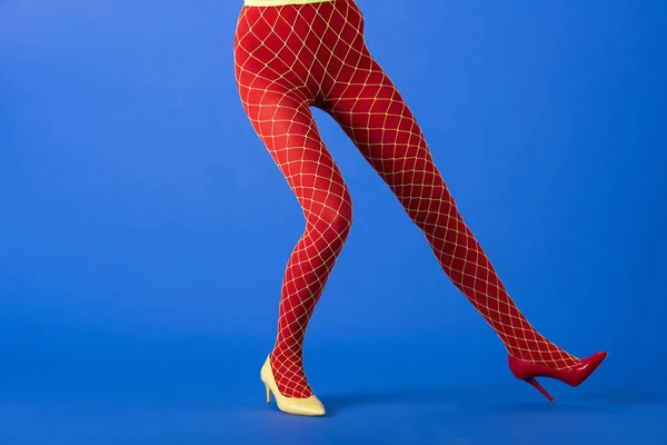 Vista recortada del modelo de moda en medias de rejilla, tacones amarillos y rojos posando en azul - foto de stock