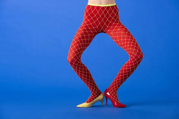 Vista recortada de modelo elegante en medias de rejilla, tacones amarillos y rojos de pie en azul - foto de stock