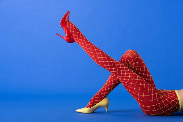 Cropped view of woman in fishnet tights, yellow and red heels lying and posing on blue — Stock Photo