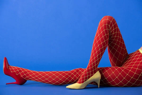 Cropped view of woman in bright fishnet tights, yellow and red heels lying on blue — Stock Photo