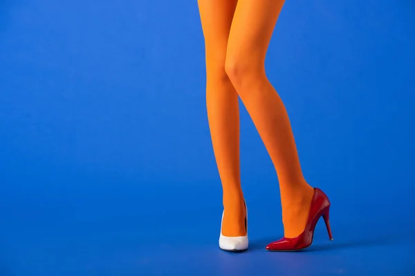 Vista recortada del modelo de moda en medias de color naranja, tacones blancos y rojos posando en azul - foto de stock