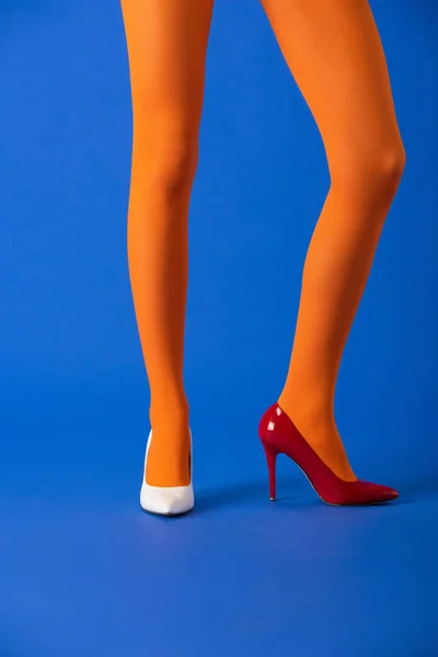 Vista recortada de modelo en medias de color naranja, tacones blancos y rojos posando en azul - foto de stock