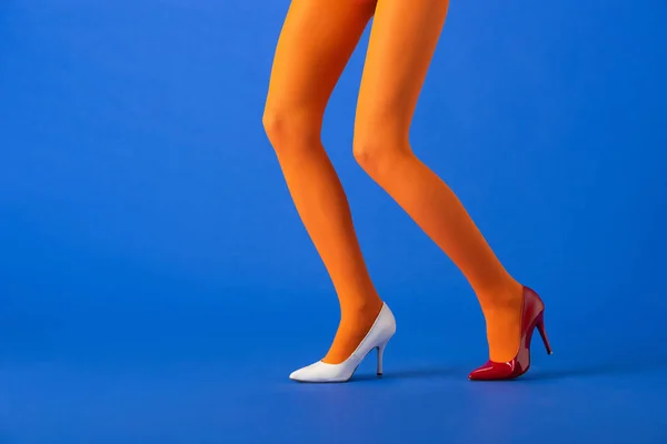 Vista recortada de modelo elegante en medias de color naranja, tacones blancos y rojos posando en azul - foto de stock