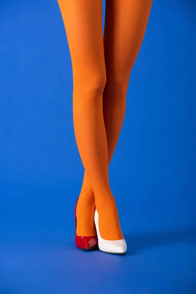 Vista recortada del modelo de moda en medias naranja, tacones blancos y rojos de pie con las piernas cruzadas en azul - foto de stock