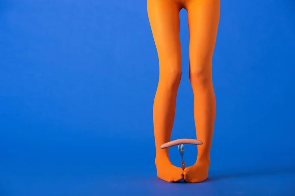 Vista recortada de la mujer en medias de color naranja brillante sosteniendo tenedor y salchicha con las piernas en azul - foto de stock