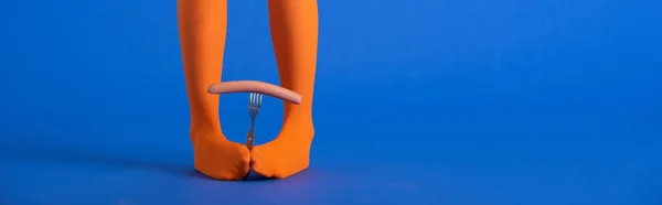 Panoramic shot of woman in bright orange tights holding fork and sausage with legs on blue — Stock Photo