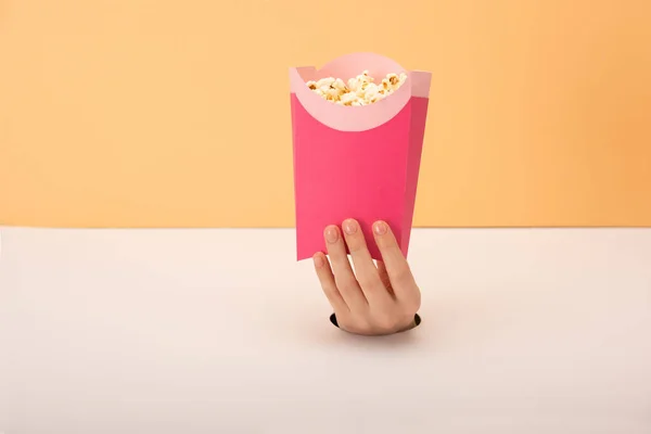 Vista ritagliata del foro con mano di donna che tiene secchio con popcorn su arancione e bianco — Foto stock