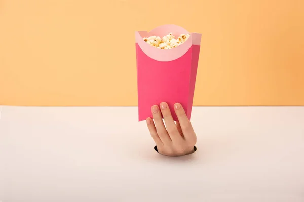 Vista recortada del agujero con mano de mujer sosteniendo cubo con palomitas de maíz en naranja y blanco - foto de stock