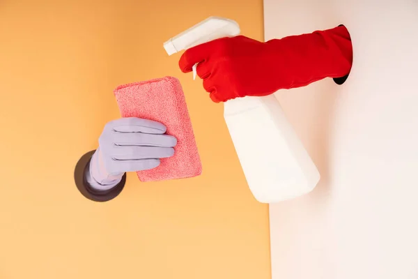 Vue recadrée des mains dans des gants tenant le flacon pulvérisateur et l'éponge sur orange et blanc — Photo de stock