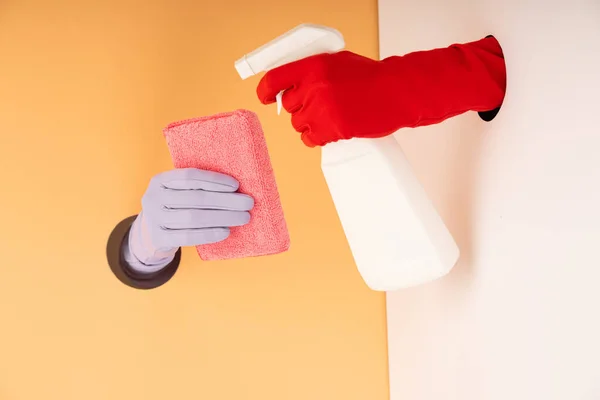 Vista recortada de las manos en guantes sosteniendo botella de spray y esponja en naranja y blanco - foto de stock