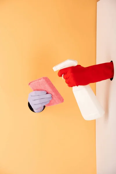 Vista recortada de las manos en guantes sosteniendo botella de spray y esponja en blanco y naranja - foto de stock