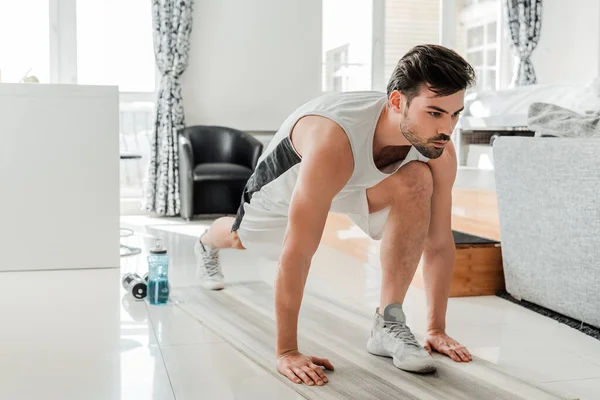 Handsome man training on fitness mat near barbells and sports bottle at home — Stock Photo