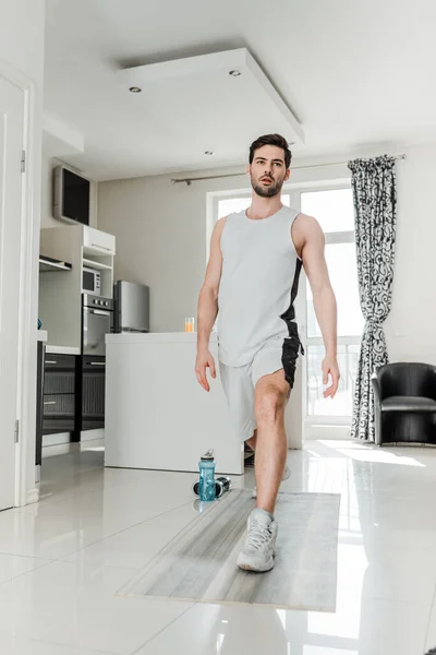Schöner Mann trainiert auf Fitnessmatte in der Nähe von Sportflasche und Langhanteln zu Hause — Stockfoto